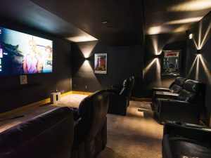 Dimly lit home theater with black leather reclining seats facing a large screen showing a movie poster scene.