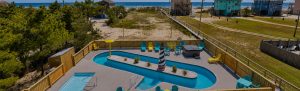 riohead | Swack Vacations Aerial view of a beachside pool deck with a curved pool, a central island feature resembling a lighthouse, and colorful lounge chairs nearby.