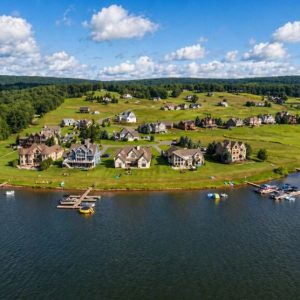 Aerial view of a lakeside neighborhood with houses along a grassy shoreline, docks, and boats on calm water.