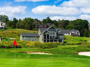 tc-Section-1 | Swack Vacations Golf green with a red flag, a sand bunker nearby, and a row of houses on a hill under a blue sky with clouds.