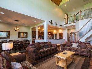 Spacious living room with brown leather sofas, a wooden coffee table, and a separate dining area; open kitchen in the background and a staircase leading to a balcony loft.
