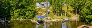 Lakeside yard with a winding path through tall trees leading to a blue-roof gazebo over a dock on calm water, with houses in the background.