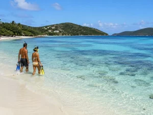 A couple carrying masks and fins enters the calm, clear water at Sapphire Beach with reef patches visible below.