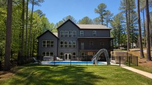 Two-story gray house with large windows overlooking a fenced-in backyard pool and a curved slide, set among tall trees under a clear blue sky.