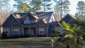 Front view of a blue house with stone veneer, gabled roof, and a green lawn surrounded by trees.
