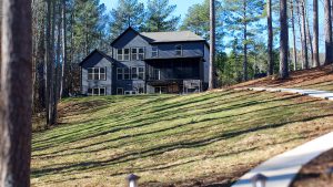 Two-story dark-blue house with many windows, screened porch, nestled among pine trees on a grassy slope; curved white fence in the foreground.