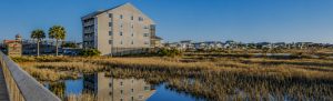 Row of beachside condos by a marsh, with a wooden boardwalk on the left and calm water reflecting the buildings.