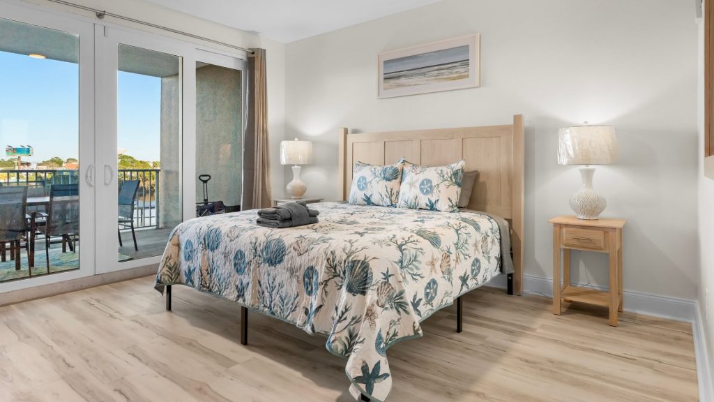 Bright bedroom with a light wood bed, ocean-themed quilt and pillows, two matching nightstands with lamps, and sliding glass doors to a balcony.