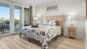 mm-up 1 | Swack Vacations Bright bedroom with a light wood bed, ocean-themed quilt and pillows, two matching nightstands with lamps, and sliding glass doors to a balcony.