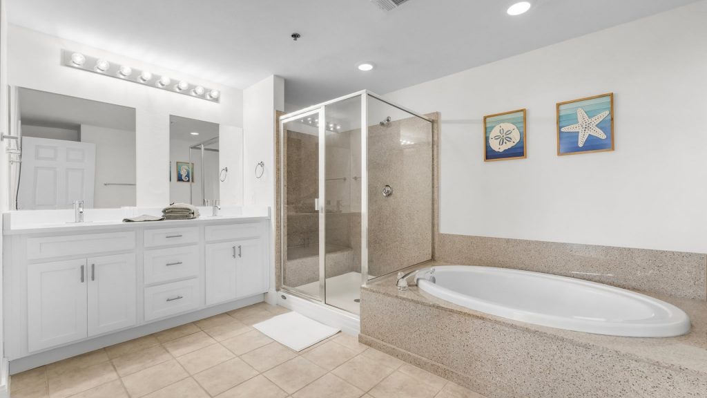 Bright bathroom with a white double-sink vanity, glass-enclosed shower, and a built-in soaking tub; beige tiles and beach-themed artwork along the wall.