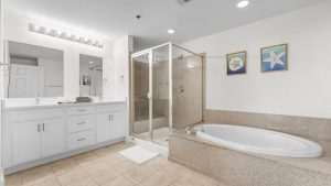 Bright bathroom with a white double-sink vanity, glass-enclosed shower, and a built-in soaking tub; beige tiles and beach-themed artwork along the wall.
