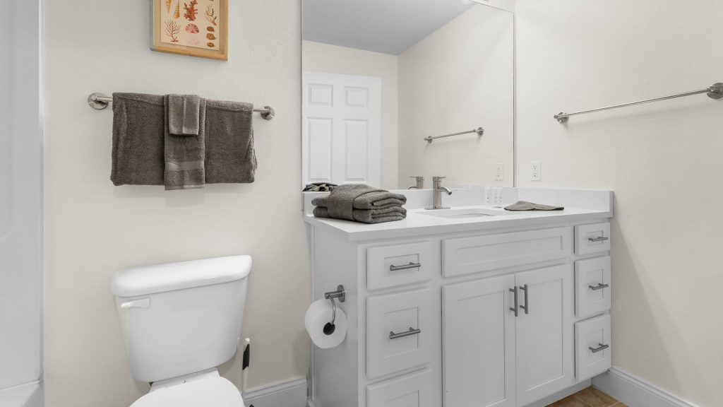 White bathroom with a single-sink vanity, gray towels on a metal bar, and a large wall mirror over the sink office-style.