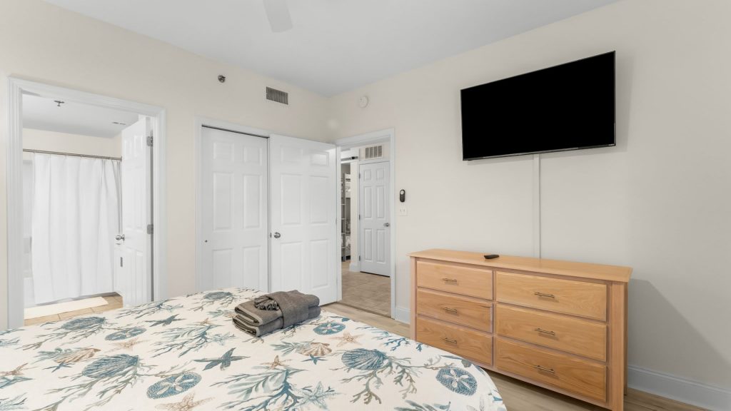 Bedroom with a sea-life patterned quilted bed, folded towels on top, and a wooden dresser beneath a wall-mounted TV.