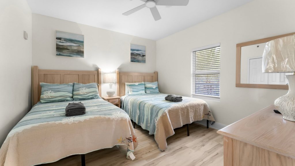 Two twin beds with ocean-blue bedspreads in a bright coastal hotel room, with a shared nightstand and lamp between them.