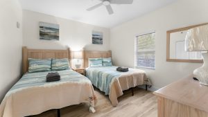mm-up 5 | Swack Vacations Two twin beds with ocean-blue bedspreads in a bright coastal hotel room, with a shared nightstand and lamp between them.