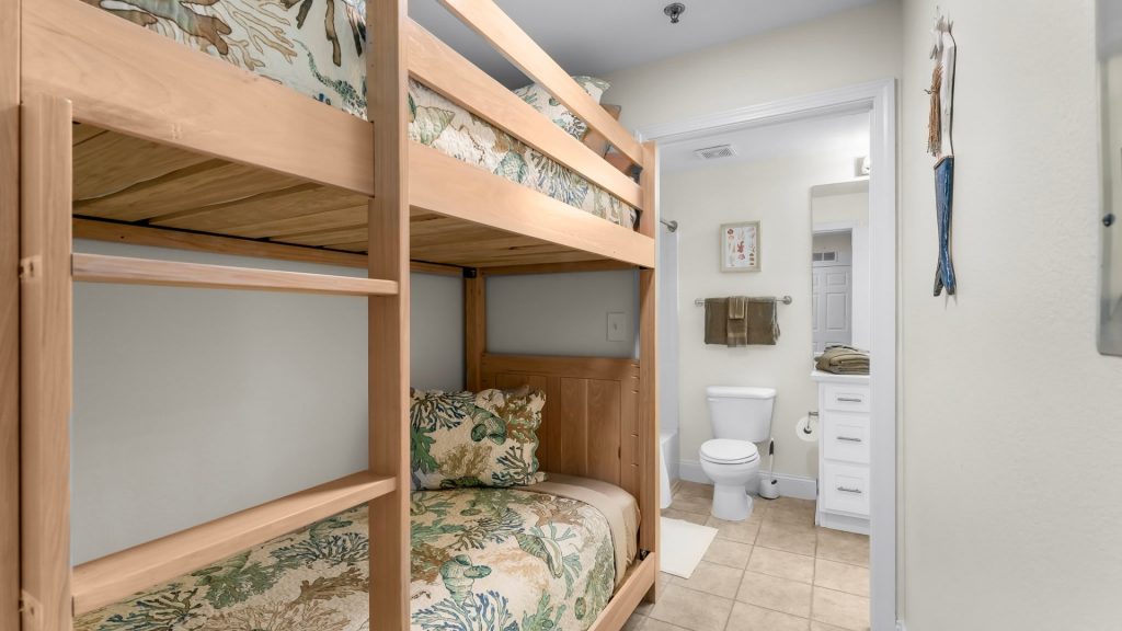 Wooden twin-over-twin bunk bed with tropical bedding in a compact bedroom, opening to a bathroom with toilet and sink visible.