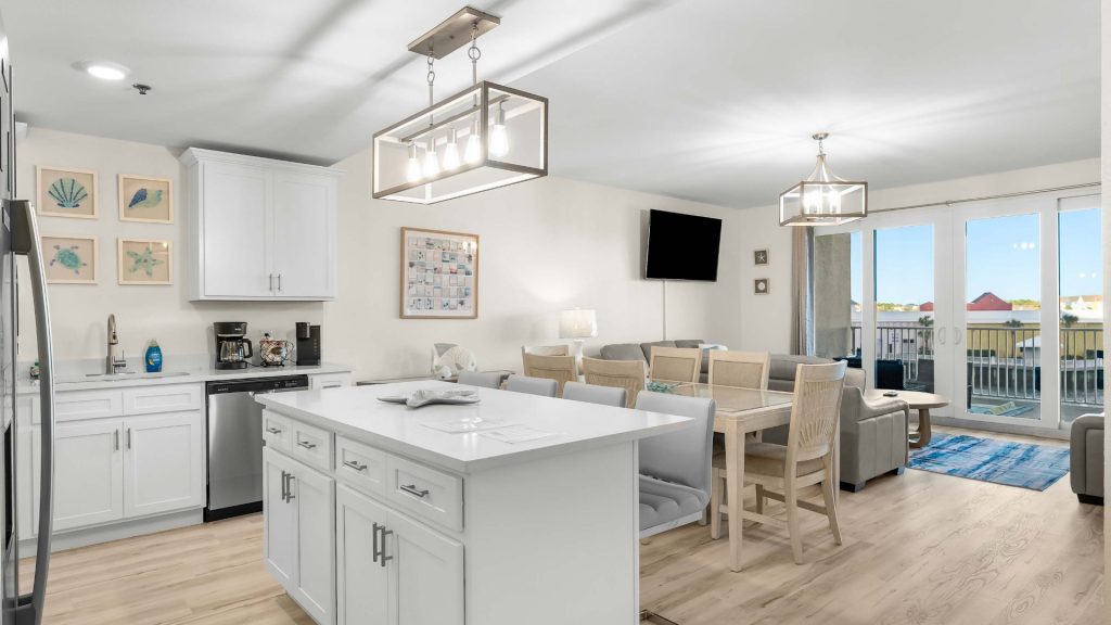 Bright open-plan kitchen, dining, and living area with white cabinets, island, and large windows to a balcony view overlooking the coast/Home by beach.