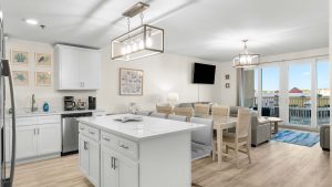 Bright open-plan kitchen, dining, and living area with white cabinets, island, and large windows to a balcony view overlooking the coast/Home by beach.