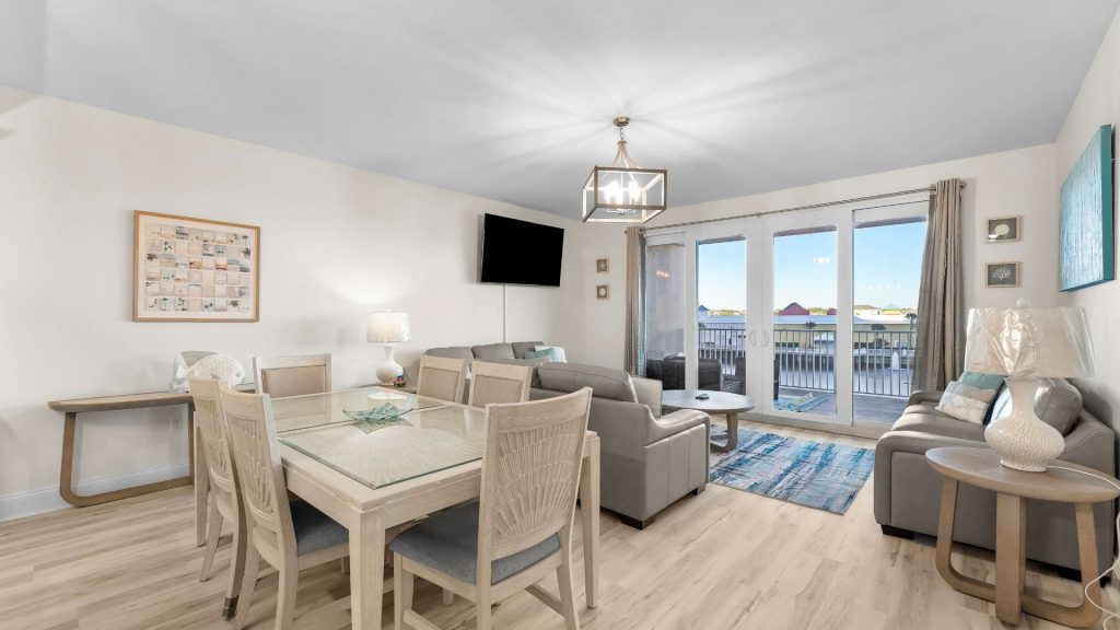 Bright coastal living space with a glass dining table, beige chairs, a gray sectional, and a balcony door overlooking water.