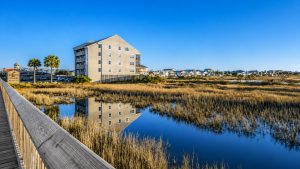 murrells-inlet-exterior-up | Swack Vacations Boardwalk along a marsh with a beige apartment building and blue sky, water reflecting the scene above.