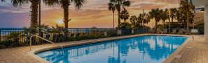 Swimming pool at sunset with palm trees, ocean view, and lounge chairs along a fenced pool deck by the sea