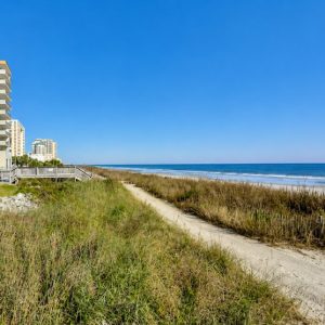 Path through sandy dunes toward the blue sea, with buildings visible to the left under a clear sky.