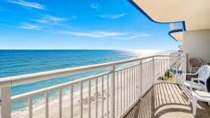 Balcony view of a blue ocean and sandy beach under a clear sky, with white chairs and railing in the foreground.