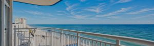 Balcony with white chair overlooking a sandy beach and blue ocean under a bright sky.