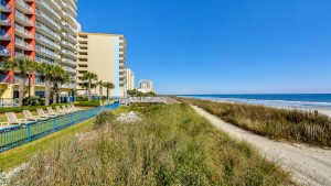 Hotel buildings line a sunny sandy beach with palm trees and a wooden boardwalk to the ocean, under a blue sky, left side of the image.