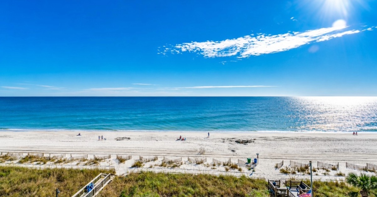 Sunny beach scene with white sand, turquoise water, and a few people relaxing along the shore; a boardwalk runs in the foreground.