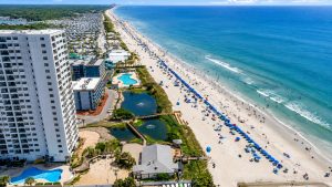 Aerial view of a crowded beach resort with high-rise buildings, rows of umbrellas, and turquoise ocean along white sand.