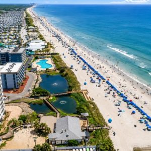 toes-in-sand-section-2 | Swack Vacations Aerial view of a crowded beach with blue umbrellas along the white sand, hotel buildings, and pool areas nearby nearby.