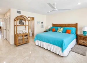 Hotel bedroom with a turquoise bedspread, white skirt, and tropical throw pillows on a wicker headboard bed, flanked by matching nightstands and lamps.