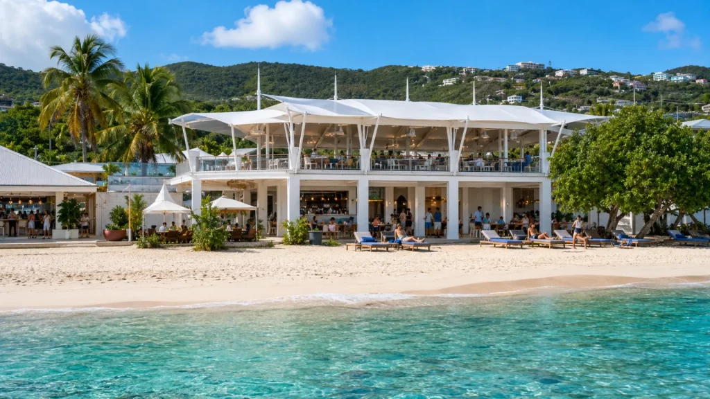 Open-air two-story beachfront pavilion with bar seating, lounge chairs, white sand, palm trees, and turquoise Caribbean water.