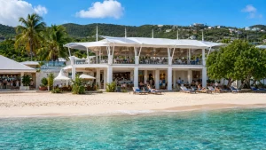 Open-air two-story beachfront pavilion with bar seating, lounge chairs, white sand, palm trees, and turquoise Caribbean water.