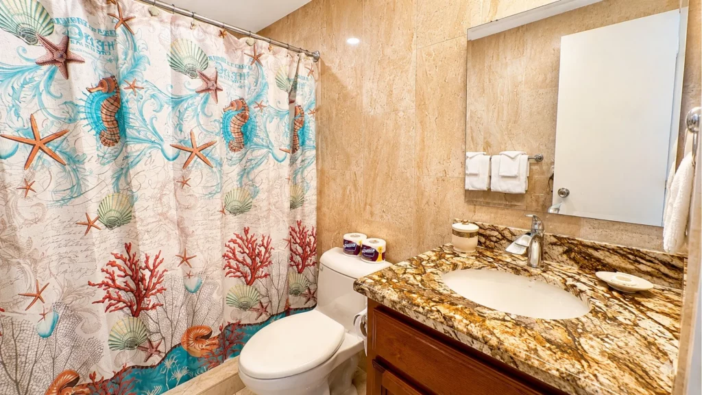 Bathroom with an ocean-themed shower curtain, beige marble walls, and a brown granite vanity top with sink.