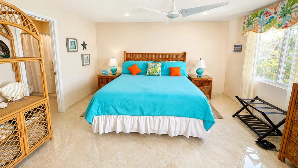 Bright tropical bedroom with a wicker headboard king bed in turquoise bedding and orange/patterned pillows, side tables, ceiling fan, and sunny window.
