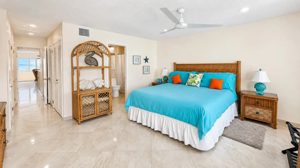Bright hotel bedroom with turquoise bedding, orange throw pillows, and a wicker headboard flanked by matching nightstands with teal lamps under a ceiling fan.
