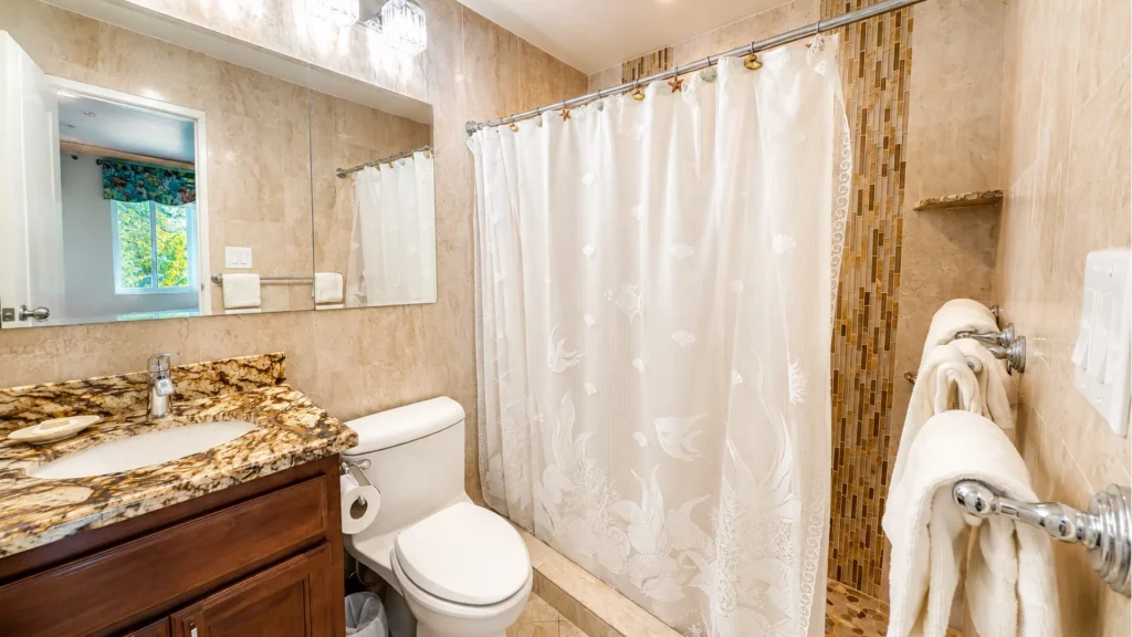 Beige tiled bathroom with a granite countertop vanity, toilet, and a white patterned shower curtain; towels on a rack nearby.