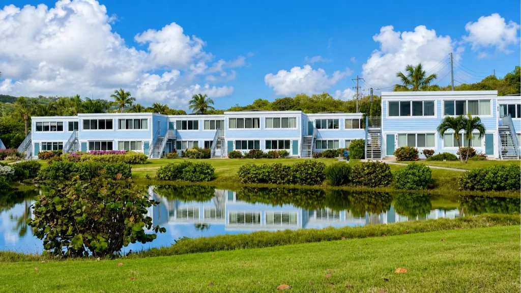 Row of light-blue, two-story coastal apartment buildings lining a calm water body with palm trees and green lawn in front.