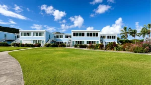 Blue two-story apartment buildings with white trim lining a landscaped lawn under a bright blue sky; walkways and stairs lead to entrances, palm trees in the background