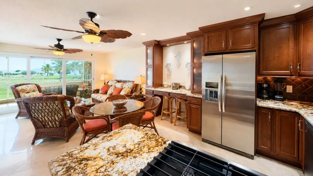 Open-plan kitchen and living room with dark wood cabinets, granite countertops, stainless fridge, and wicker seating, looking out to a lawn with palm trees through large windows.