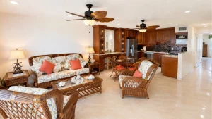 crystal-cove-living-space-1 | Swack Vacations Bright tropical living room with wicker sofas and floral cushions, opening to a dark-wood kitchen.