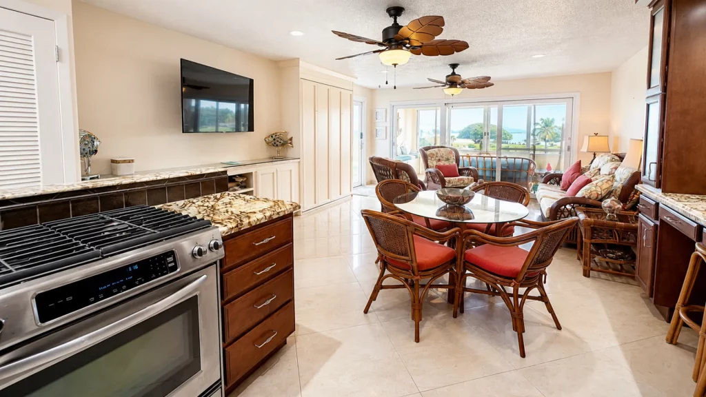 Bright open-plan kitchen, dining, and living area with a glass dining table, wicker chairs, and a wall-mounted TV on beige walls.