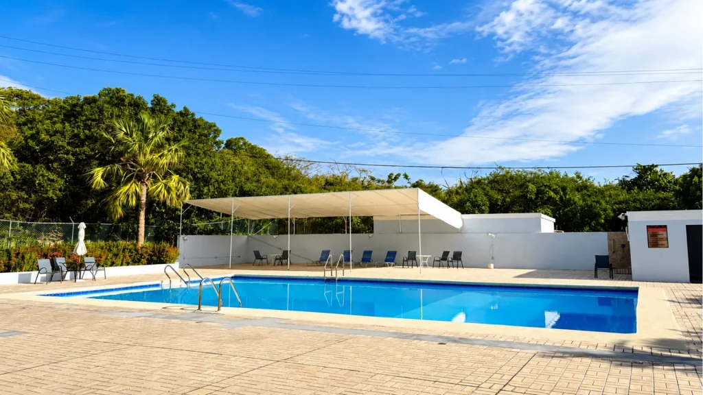 Poolside outdoor area with a bright blue pool, tan tiled patio, and a white canopy over several lounge chairs under a clear sky.