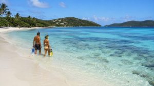 A couple carrying masks and fins enters the calm, clear water at Sapphire Beach with reef patches visible below.