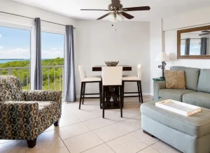 Bright living room with ocean view through glass doors, a teal sofa, patterned armchair, and a dark wood dining table with four cream chairs.