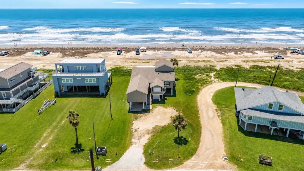 Aerial view of beach houses along a sandy shoreline with the ocean beyond and green lawns in the foreground, on a sunny day.4fa