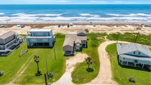 Aerial view of beach houses along a sandy shoreline with the ocean beyond and green lawns in the foreground, on a sunny day.4fa