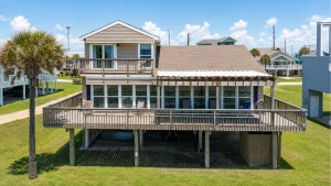 Coastal two-story house on stilts with a large wraparound deck and sliding glass doors, palm tree in the foreground under a blue sky.